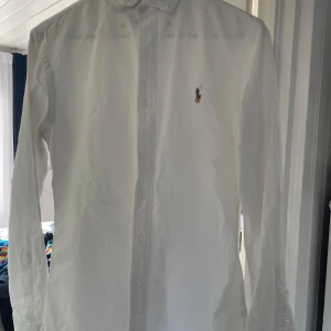 Vit skjorta från Ralph Lauren - Säljer en klassisk vit skjorta från Ralph Lauren med broderad logotyp på bröstet. Skjortan har långa ärmar och knappar framtill. Perfekt för en stilren look.
