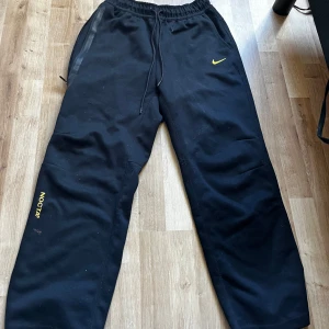 Svarta mjukisbyxor från Nike NOCTA - Svarta mjukisbyxor från Nike NOCTA med gul logga och textdetaljer. Byxorna har en elastisk midja med snörning och sidofickor. Perfekta för en avslappnad stil.