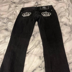 Svarta jeans från Victoria Beckham - Snygga svarta jeans från Victoria Beckham med broderade kronor på bakfickorna. Jeansen har en klassisk design med knapp och dragkedja framtill. Perfekta för en stilren look. Byxorna är low waist och är i storleken 34. 