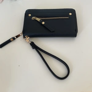 Svart plånbok med dragkedja - Snygg svart plånbok med dragkedja och avtagbar handledsrem. Plånboken har flera fack för kort och sedlar, samt en yttre ficka med dragkedja för småsaker. Perfekt för att hålla ordning på dina tillhörigheter.