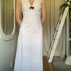 Vit ärmlös maxiklänning med broderi - Elegant vit långklänning med v-ringning och en söt svart rosett framtill. Klänningen är ärmlös och har ett vackert broderat blommönster. Perfekt för sommaren eller studenten! Den är ny med prislapp kvar!!