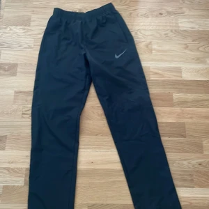 Nike byxor - Säljer ett par svarta träningsbyxor från Nike med Dri-FIT-teknologi. Byxorna har en elastisk midja och en diskret Nike-logga på benet. Perfekta för träning och aktivitet.