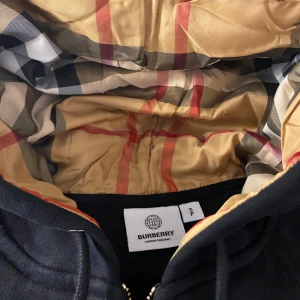 Svart hoodie från Burberry - Säljer en stilren svart hoodie från Burberry med klassiskt rutigt foder i huvan. Jackan har dragkedja framtill och ribbade muddar vid ärmslut och nederkant. Perfekt för en casual look.