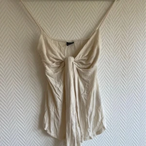 Beige linne med knyt - Säljer ett stilrent beige linne med knyt framtill. Toppen har tunna axelband och en luftig passform, perfekt för varma dagar. Enkelt att matcha med olika outfits. Har lappar kvar 