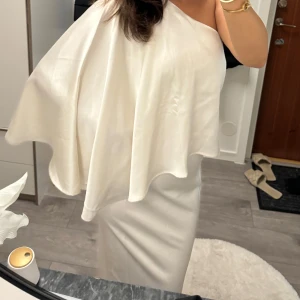 Vit axelbandslös klänning - Elegant vit axelbandslös klänning med asymmetrisk design. Tillverkad i ett glansigt material som ger en lyxig känsla. Perfekt för speciella tillfällen. Med prislapp 