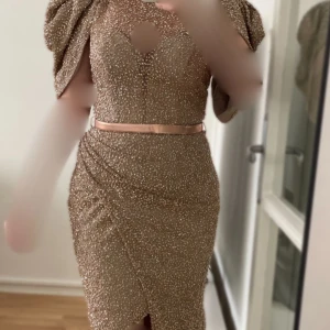 Lyxig glittrig guldfärgad klänning- använd en gång, i toppskick - Säljer en elegant guldfärgad festklänning i lyxigt glittrande tyg. Endast använd i 6,7 timmar.  STORLEK 36 Slits framtill och vackra axeldetaljer.  Ett midjebälte medföljer och kan användas om man vill eller tas bort för en annan look Nypris 3500 kr säljer för 2000. Fler bilder kan skickas vid intresse. Hör av dig om du har frågor