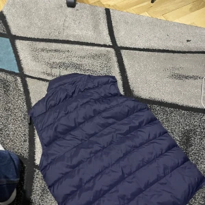 Mörkblå dunväst från Moncler - Snygg mörkblå dunväst från Moncler med dragkedja och två fickor framtill. Perfekt för kyligare dagar och ger en stilren look. Västen har en hög krage och Moncler-loggan på bröstet.