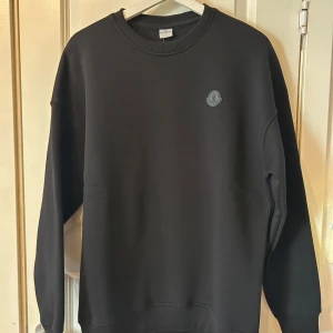 Svart sweatshirt från Moncler - Säljer en stilren svart sweatshirt från Moncler med rund hals och ribbade muddar. Tröjan har en diskret Moncler-logga på bröstet. Perfekt för en avslappnad och trendig look.