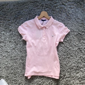 Ljusrosa pikétröja gant - Säljer en ljusrosa pikétröja med krage och knappar framtill. Tröjan har korta ärmar och en liten logga på bröstet. 