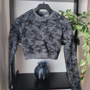 Svart sporttopp från Gymshark i storlek S - En kamouflage träningstopp från märket Gymshark. Storlek S. Knappt använd då jag insett att jag inte trivs i denna typen av topp. Snyggt inbroderad Gymsharklogga på ena sidan. Toppen har tummhål. Även snygga detaljer på ryggen i form av ovala utskärningar. Stretchig i materialet. Logga på ena ärmen. Liten detalj mellan bysten.