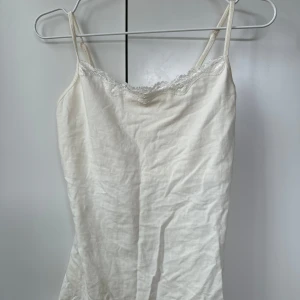 Vit linne med spetsdetaljer - Säljer ett stilrent vitt linne med smala axelband och spetsdetaljer vid halsringningen. Perfekt för varma sommardagar eller som ett basplagg i garderoben. Den har även en inbyggd topp