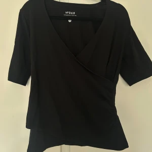 Svart omlottopp från No365 - Snygg svart omlottopp från No365 i storlek M med korta ärmar. Perfekt för en stilren look. Toppen har en v-ringad design som ger en elegant touch. Väldigt fin passform! 🩷