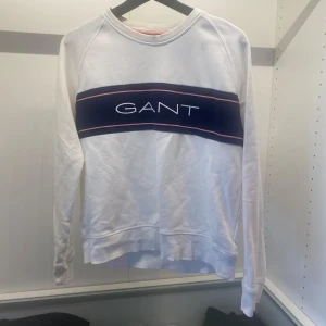 Vit sweatshirt från GANT - Säljer en stilren vit sweatshirt från GANT med ett blått och rosa band över bröstet där märkesnamnet är tryckt. Tröjan har långa ärmar och en rund halsringning. 