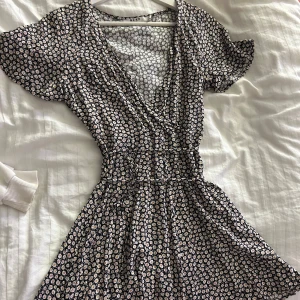 Blommig dress(shorts) från NA-KD - Säljer en söt blommig dress(shorts istället för kjol) från NA-KD i storlek 34. Klänningen har en omlott-design med korta ärmar och ett fint blommönster i vitt och gult på svart bakgrund. Perfekt för sommarens alla tillfällen! 🌼
