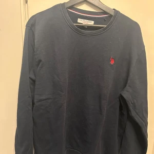 Mörkblå tröja från U.S. Polo Assn. - Säljer en stilren mörkblå tröja från U.S. Polo Assn. med ett rött broderat emblem på bröstet. Tröjan har långa ärmar och rund halsringning, perfekt för en avslappnad look.
