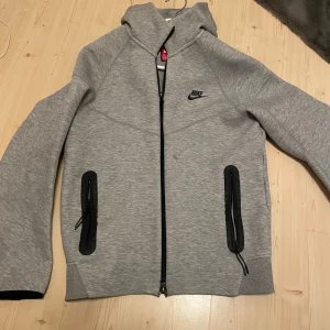 Nike tech fleece - Säljer en grå hoodie från Nike med dragkedja och huva. Jackan har svarta detaljer vid fickorna och en liten Nike-logga på bröstet. Perfekt för en sportig look eller avslappnade dagar.