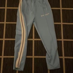 Blå träningsbyxor från Palm Angels S/M - Snygga ljusblå träningsbyxor från Palm Angels med vita och svarta ränder längs sidorna. Byxorna har elastisk midja med snörning och dragkedjor vid bensluten för en justerbar passform. Säljer dom då jag ej har användning till dom mer priset kan diskuteras tar trades .