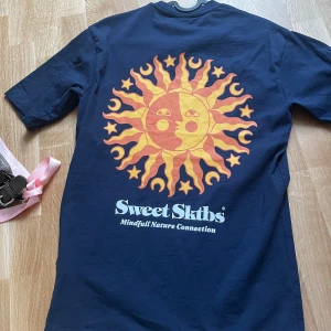Mörkblå t-shirt från Sweet Skibs - Säljer en mörkblå t-shirt från Sweet Skibs med ett stort sol- och månmotiv i orange och gult på ryggen. T-shirten har korta ärmar och rund hals. Perfekt för dig som gillar unika tryck och skön stil. 🌞🌙