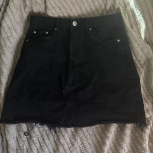 Svart jeanskjol - Snygg svart jeanskjol med rå kant och klassisk femficksdesign. Perfekt för en avslappnad stil. Kjolen har en knapp och dragkedja framtill.