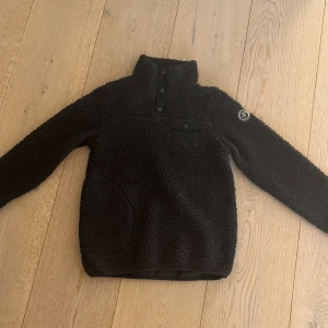 Svart/mörkblå fleece tröja från Pelle P - Mysig svart/mörkblå fleece tröja från Pelle P med hög krage och knappar framtill. Tröjan har en praktisk bröstficka med knapp och en rund logga på ärmen. Perfekt för kyliga dagar!