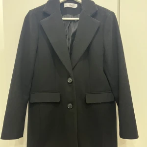 Svart kappa  - Elegant svart kappa från Ellos med klassisk design och knappar framtill. Perfekt för kyligare dagar med sin stilrena look och långa ärmar.