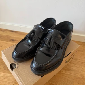 Svarta loafers med tofsar från Dr. Martens - Säljer ett par klassiska svarta loafers från Dr. Martens med dekorativa tofsar och flätad detalj runt tån. Skorna har en rund tå och platt sula, tillverkade i blankt skinn. Perfekta för dig som vill ha en stilren och tidlös look. Postas i sin original förpackning enligt bilden ovan.   Endast testade utav mig, och använda ett fåtal gånger. från tidigare ägare. Alltså väldigt mycket som nya. 😊 Nypris 2000kr