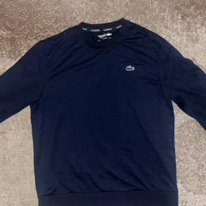 Mörkblå långärmad tröja från Lacoste Sport - Säljer en mörkblå långärmad tröja från Lacoste Sport med klassisk passform. Tröjan har det ikoniska krokodilmärket på bröstet och en sportig detalj i grönt och gult längs sidan. Perfekt för en sportig och stilren look.