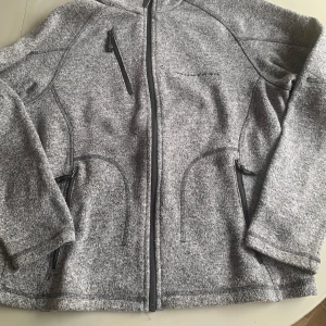 Grå fleecejacka från Ford - Mysig grå fleecejacka från Ford med hel dragkedja framtill och tre fickor med dragkedja. Perfekt för lager-på-lager och svalare dagar. Enkel och stilren design med diskret logga på bröstet.
