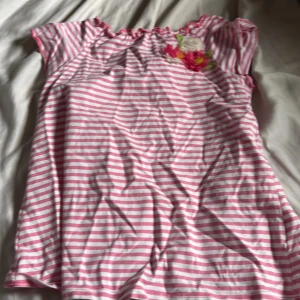 Rosa och vit randig topp med blommor - Söt topp med rosa och vita ränder och små blommor broderade vid halsen. Kortärmad modell med lätt rynkad halsringning. Perfekt för en somrig look!