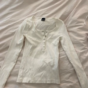 Vit ribbad långärmad topp från Gina Tricot - Säljer en vit ribbad långärmad topp från Gina Tricot i storlek XS. Toppen har knappar framtill och en djupare rundad halsringning. Perfekt att styla till olika outfits!