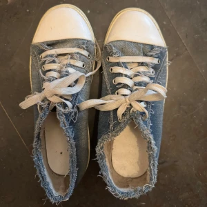 Blå jeanssneakers med fransig kant - Säljer ett par blå sneakers i jeansmaterial med vit sula och tå. Skorna har fransiga kanter runt öppningen och vita snören. Perfekta för en avslappnad och trendig stil.