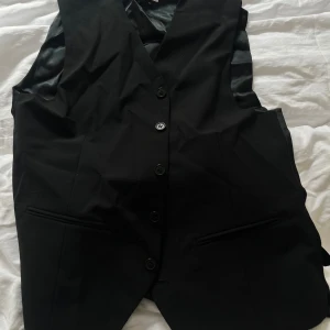 Svart kostymväst med knappar - Snygg svart väst med klassisk passform och knäppning framtill. Västen har två fickor och är fodrad med glansigt tyg på insidan. Perfekt att bära över en skjorta för en stilren look.