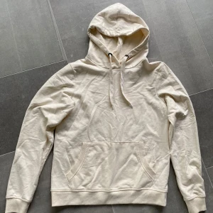 Beige hoodie med ficka - Säljer en stilren beige hoodie med huva och dragsnören. Tröjan har en stor magficka och ribbade muddar vid ärmslut och nederkant. Perfekt för en avslappnad look.
