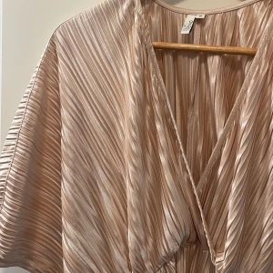 Champagne rosa plisserad klänning från NLY Trend - Säljer en champagne rosa plisserad klänning från NLY Trend. Klänningen har omlott-skärning, v-ringning och vida korta ärmar. Materialet är glansigt och ger en lyxig känsla, perfekt för speciella tillfällen. Super skön dessutom.