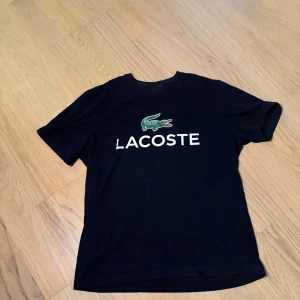 Svart t-shirt från Lacoste med logga - Svart t-shirt från Lacoste med klassisk krokodil-logga och vit text på bröstet. Regular fit och rund halsringning. Perfekt för en stilren och enkel look.