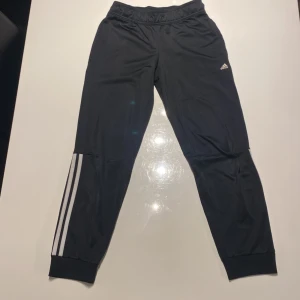 Svarta träningsbyxor från Adidas med vita ränder - Svarta Adidas träningsbyxor med klassiska vita ränder längs benen och liten Adidas-logga på låret. Byxorna har resår i midjan och smal passform vid anklarna. Perfekta för sport eller chill. I storlek 152 (11-12 år). Ett jättelitet defekt som knappt går och synas. För ENDAST 50kr så får du dessa extremt bra svarta Adidas träningsbyxor!