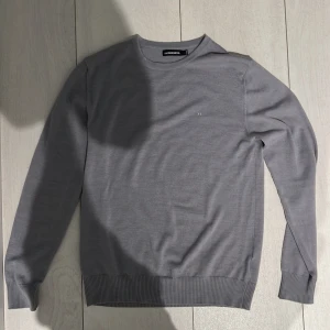 Grå tröja från J.Lindeberg - Säljer en stilren grå tröja från J.Lindeberg i storlek L. Tröjan passar M,Köpt på NK diskret logga på bröstet. Perfekt nu infor sommaren med att  Materialet känns mjukt och lätt samt luftigt.