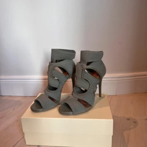 Gråa / kahki, klackar med öppen tå - Säljer ett par klackar med hög klack och öppen tå. Skorna har ett unikt mönstrat material och breda remmar som ger en cool look. Perfekta för dig som vill sticka ut med snygga detaljer och modern design.