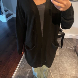 Svart stickad kofta från 157 - Mysig svart stickad kofta från 157 med två fickor framtill. Koftan har lång ärm och en avslappnad passform, perfekt att slänga på sig över en topp. Enkel och stilren design.