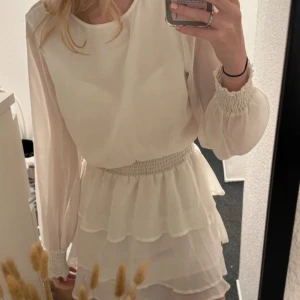 Vit volangklänning från Gina Tricot - Säljer en söt vit kortklänning från Gina Tricot med långa transparenta ärmar, smock i midjan och flera lager volanger i kjolen. Klänningen har en öppen detalj i ryggen och är perfekt för somriga tillfällen.