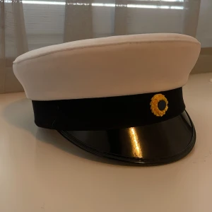 Studentmössa med svart skärm och gulddetalj - Klassisk studentmössa med vit ovandel, svart band och blank svart skärm. Framtill finns en gulddetalj. Insidan har ett foder i blått och gult satin. Perfekt för studenten!