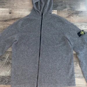 Grå stickad hoodie från Stone Island - Säljer en grå stickad hoodie från Stone Island med dragkedja framtill och klassisk logotyp på ärmen. Tröjan har huva och långa ärmar, perfekt för kyligare dagar. Enkel och stilren design som passar till mycket.