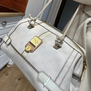 Vit handväska från Guess - Säljer en stilren vit handväska från Guess med diskret mönster och gulddetaljer. Väskan har dragkedja upptill, dubbla handtag och dekorativa spännen på sidorna. Perfekt för dig som gillar klassisk och elegant design.