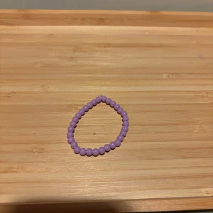 Lila pärlarmband - Säljer ett enkelt och stilrent armband med lila pärlor. Perfekt för att addera lite färg till din outfit. Elastiskt band gör det lätt att ta på och av.
