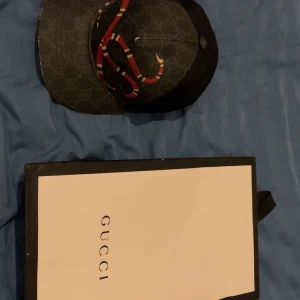 Gucci Kingsnake keps - Säljer en svart keps från Gucci med diskret GG-mönster och en stor röd och vit orm på framsidan. Kepsen har justerbart band bak med röd och blå rand. Perfekt för dig som vill sticka ut med en lyxig accessoar.