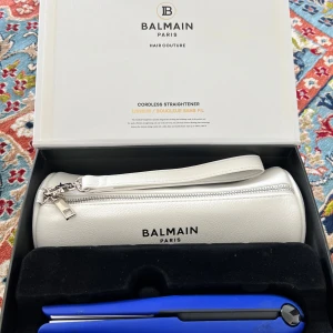 Balmain Paris sladdlös plattång med vit necessär - Exklusiv sladdlös plattång från Balmain Paris i matt blå finish med silvriga plattor. Kommer med en stilren vit necessär i läderimitation med dragkedja och Balmain-logga. Perfekt för styling on-the-go och enkel att ta med.