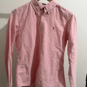 Rosa skjorta från Ralph Lauren - Säljer en klassisk rosa skjorta från Ralph Lauren i skinny fit-modell. Skjortan har knappar framtill, button-down krage och den ikoniska broderade loggan på bröstet. Perfekt för dig som gillar stilrena och tidlösa plagg.