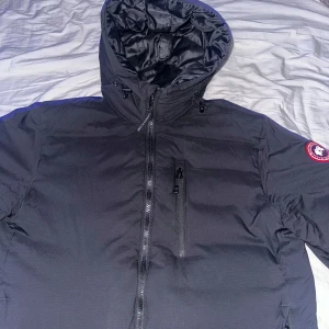Svart dunjacka från Canada Goose - Säljer en svart dunjacka från Canada Goose med huva och dragkedja framtill. Jackan har en bröstficka med dragkedja och klassisk logotyp på ärmen. Perfekt för kalla dagar och riktigt varm.