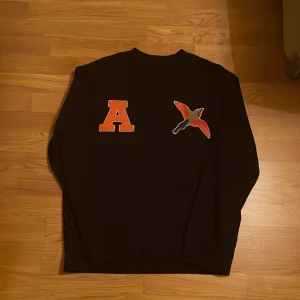 Svart sweatshirt från Axel Arigato med broderad fågel - Svart sweatshirt från Axel Arigato med en stor orange bokstav 'A' och en broderad fågel i flera färger på bröstet. Tröjan har rund hals och långa ärmar. Perfekt för dig som gillar streetwear och snygga detaljer.