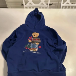 Blå hoodie med Polo Bear från Ralph Lauren - Säljer en blå hoodie från Ralph Lauren med den klassiska Polo Bear-trycket framtill. Tröjan har huva och lång ärm, perfekt för en avslappnad stil. Mysig och snygg detalj med björnen som håller julklappar. Nypris är 3000 ungefär passar bra till xs till s kanske till M
